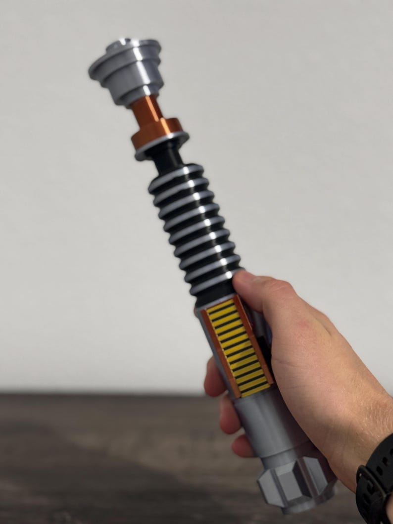 Puede incluir: Un sable de luz plateado y negro con detalles en naranja y amarillo. El accesorio tiene una secci&oacute;n negra acanalada, un mango plateado y una parte superior detallada. El sable de luz se sostiene en una mano.