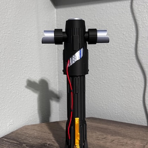 May include: A black lightsaber with a crossguard design. The hilt has a textured grip, red wiring, and a gold-coloured rectangular element. The crossguard blades are silver. The lightsaber is displayed on a wooden surface.