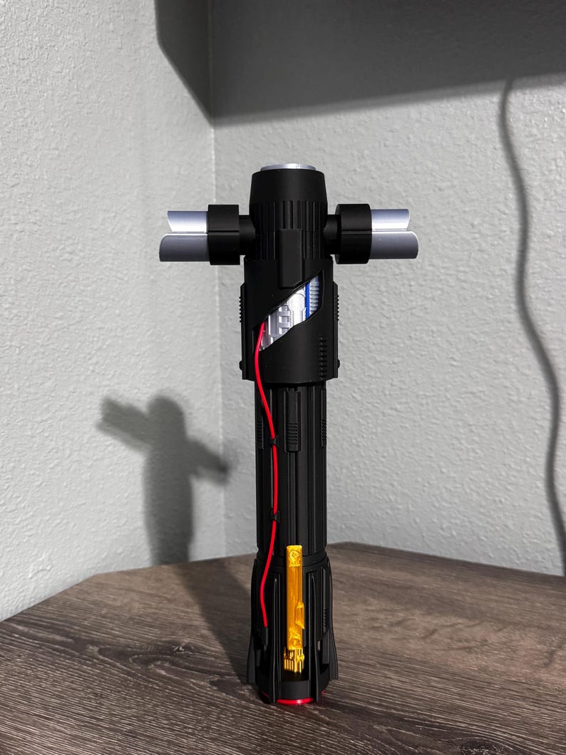 May include: A black lightsaber with a crossguard design. The hilt has a textured grip, red wiring, and a gold-coloured rectangular element. The crossguard blades are silver. The lightsaber is displayed on a wooden surface.