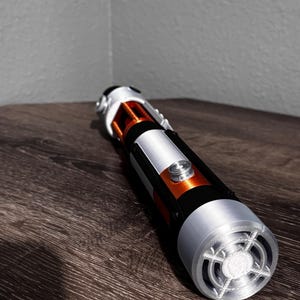 May include: A lightsaber prop in silver, black, and orange. The hilt has a silver emitter, a black grip, and orange details. The lightsaber is placed on a dark wood surface.