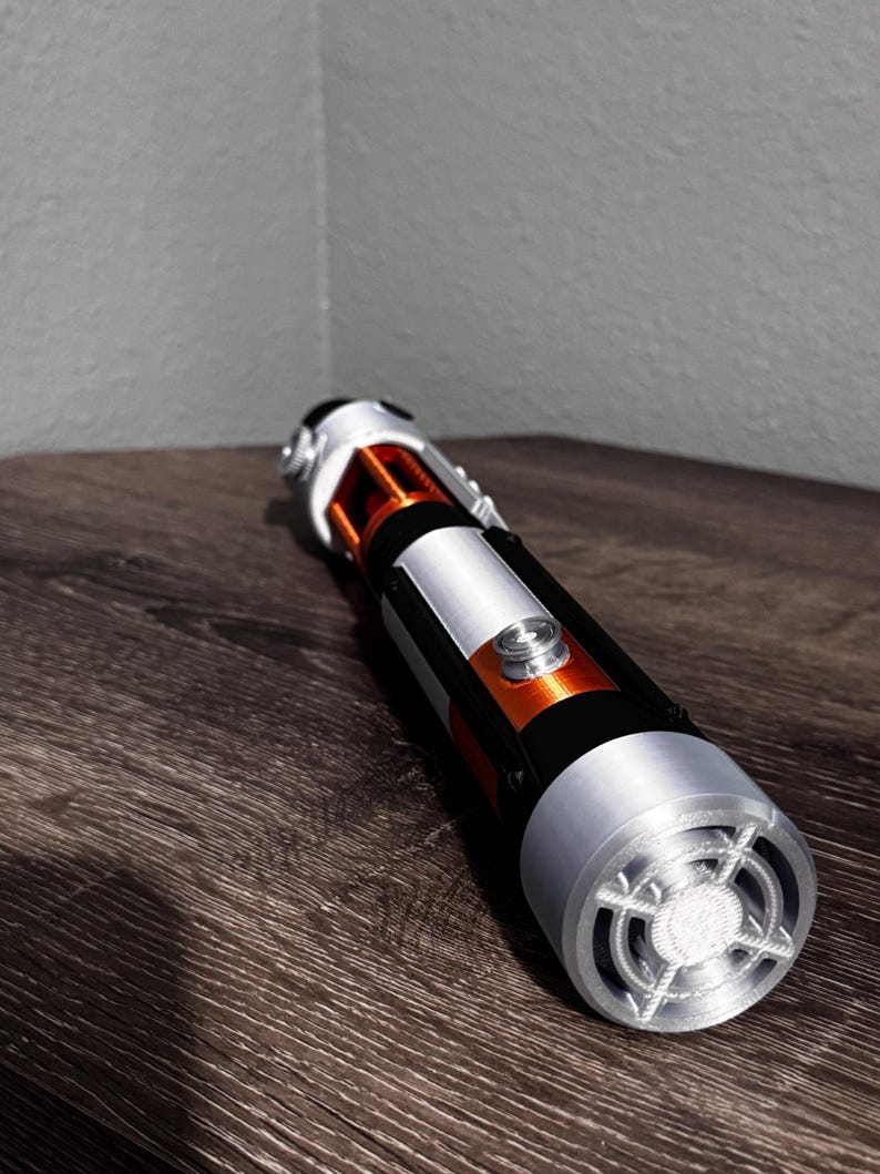 May include: A lightsaber prop in silver, black, and orange. The hilt has a silver emitter, a black grip, and orange details. The lightsaber is placed on a dark wood surface.