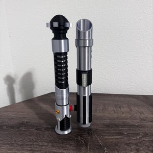 May include: Two metallic lightsabers with black and silver accents. One has a black handle with a ribbed design, while the other has a sleek, cylindrical design with a black grip and a series of lights. These props are likely for cosplay or display.