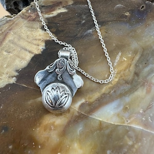 May include: A silver pendant necklace featuring a detailed lotus flower design. The pendant has an ornate, flowing shape with a circular element at the bottom. The necklace chain is delicate and silver, complementing the pendant's design. The background is a natural, earthy tone.