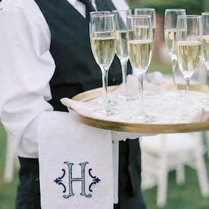 May include: A server holds a gold-rimmed tray with champagne flutes filled with sparkling wine. A white linen napkin with a blue embroidered letter "H" is draped over the server's arm. The server wears a black vest, white shirt, and black tie.