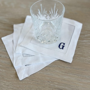 May include: A clear, patterned glass sits atop a stack of white linen coasters. The coasters have a stitched border and a navy blue embroidered letter "G" in the corner. The coasters are on a light wood surface.