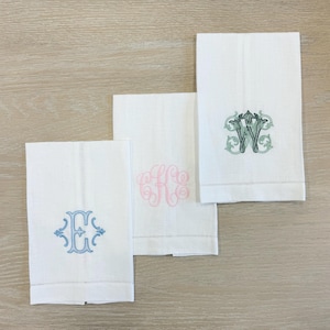 May include: Three white linen hand towels with embroidered monograms in blue, pink, and green. The towels are rectangular with a stitched border and are arranged on a light wood surface. The monograms are stylized.