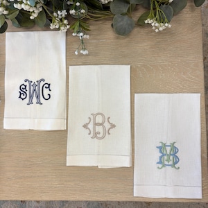 May include: Three rectangular white linen towels featuring embroidered monograms. The left towel displays a dark blue SWC monogram, the center towel features a tan B monogram, and the right towel shows a blue and green MV monogram.