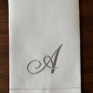 May include: A white linen hand towel with a gray embroidered letter "A". The towel has a stitched border along the bottom edge. The background is a dark wood surface.