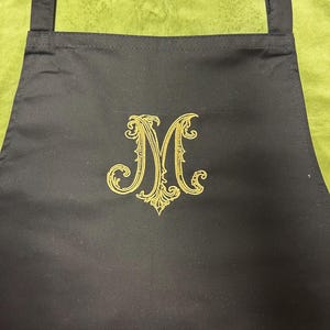 Peut inclure: Un tablier noir orn&eacute; d'une grande lettre M brod&eacute;e de couleur dor&eacute;e au centre. La lettre est con&ccedil;ue avec des volutes et des ornements complexes et tourbillonnants. Le tablier poss&egrave;de des bretelles en tissu noir aux coins sup&eacute;rieurs.