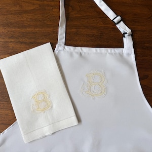 Peut inclure: Un tablier blanc et une serviette rectangulaire blanche assortie, tous deux orn&eacute;s d'une lettre B brod&eacute;e en jaune p&acirc;le dans une police cursive d&eacute;corative. Le tablier comprend une sangle de cou r&eacute;glable avec une boucle coulissante noire.