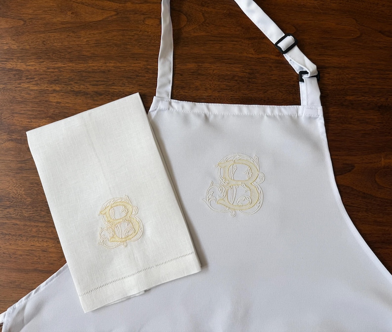 Peut inclure: Un tablier blanc et une serviette rectangulaire blanche assortie, tous deux orn&eacute;s d'une lettre B brod&eacute;e en jaune p&acirc;le dans une police cursive d&eacute;corative. Le tablier comprend une sangle de cou r&eacute;glable avec une boucle coulissante noire.