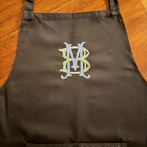 Peut inclure: Un tablier noir avec un monogramme brod&eacute; au centre. Le monogramme se compose des lettres B, M et B entrelac&eacute;es avec du fil vert clair et bleu clair. Le tablier comprend des bretelles r&eacute;glables et une section de poche sur le devant.