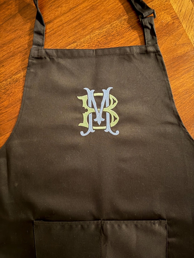 Peut inclure: Un tablier noir avec un monogramme brod&eacute; au centre. Le monogramme se compose des lettres B, M et B entrelac&eacute;es avec du fil vert clair et bleu clair. Le tablier comprend des bretelles r&eacute;glables et une section de poche sur le devant.