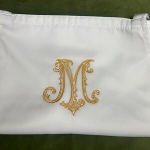 Peut inclure: Une pochette rectangulaire en tissu blanc orn&eacute;e d'une lettre M brod&eacute;e en fil dor&eacute; au centre. La pochette pr&eacute;sente une fermeture &agrave; cordon sur le dessus avec des boucles en tissu sur les c&ocirc;t&eacute;s, offrant une surface lisse et des d&eacute;tails brod&eacute;s.
