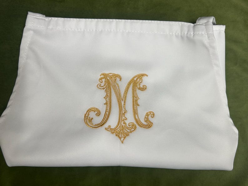 Peut inclure: Une pochette rectangulaire en tissu blanc orn&eacute;e d'une lettre M brod&eacute;e en fil dor&eacute; au centre. La pochette pr&eacute;sente une fermeture &agrave; cordon sur le dessus avec des boucles en tissu sur les c&ocirc;t&eacute;s, offrant une surface lisse et des d&eacute;tails brod&eacute;s.