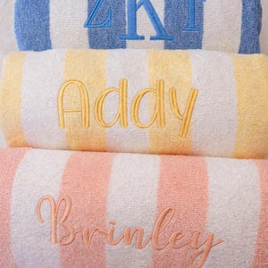 May include: Three rolled-up beach towels with embroidered names and initials. The top towel is blue and white striped with blue embroidered initials. The middle towel is yellow and white striped with the name "Addy" embroidered in gold. The bottom towel is pink and white striped with the name "Brinley" embroidered in pink.