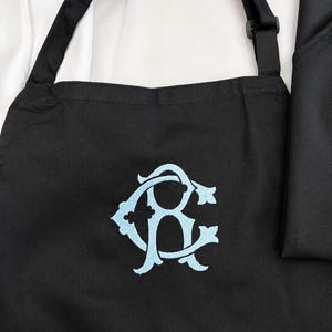 Peut inclure: Un tablier noir avec un monogramme brod&eacute; bleu clair repr&eacute;sentant les lettres C et R entrelac&eacute;es dans une police script d&eacute;corative. Le tablier comprend des bretelles noires r&eacute;glables et est plac&eacute; sur une chemise blanche et un tissu noir.