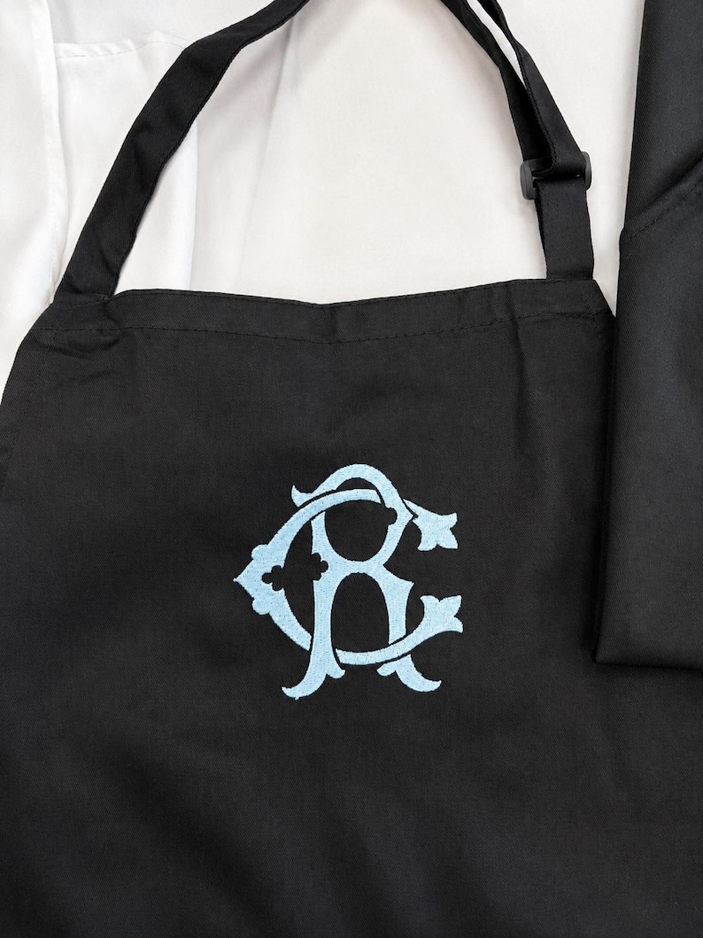 Peut inclure: Un tablier noir avec un monogramme brod&eacute; bleu clair repr&eacute;sentant les lettres C et R entrelac&eacute;es dans une police script d&eacute;corative. Le tablier comprend des bretelles noires r&eacute;glables et est plac&eacute; sur une chemise blanche et un tissu noir.
