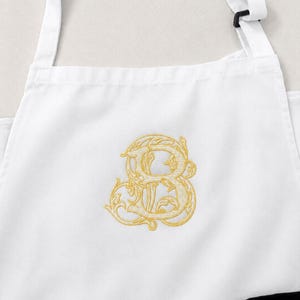 Peut inclure: Un tablier blanc orn&eacute; d'une lettre B brod&eacute;e en jaune dor&eacute; dans une police script ornementale et d&eacute;corative. Le tablier comprend des bretelles blanches avec des boucles de r&eacute;glage noires et une poche noire visible en bas &agrave; droite.