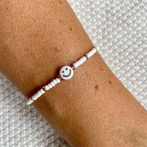 Könnte beinhalten: Weißes Perlenarmband mit einem Smiley-Anhänger. Das Armband hat kleine weiße Perlen, goldfarbene Akzente und einen zentralen Smiley-Anhänger mit gelbem Hintergrund und schwarzen Augen und Mund. Das Armband wird am Handgelenk getragen.