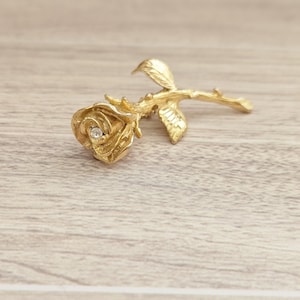 May include: A gold-toned rose brooch with a single clear rhinestone in the center of the rose. The rose is detailed with a stem and leaves.