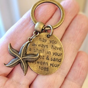 May include: A bronze-colored keychain featuring a starfish charm and a round tag. The tag is inscribed with the words "may you always have a shell in your feet & sand between your toes". The keychain has a large ring.