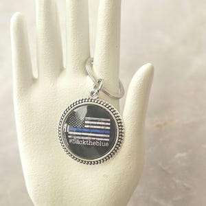 May include: A round silver keychain with a graphic of the American flag with a blue stripe, and the text "#backtheblue". The keychain is held up by a white hand.