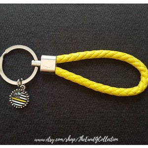 May include: A yellow braided leather keychain with a silver key ring and a round charm. The charm features a black and white American flag design with a yellow stripe. The keychain is on a dark background.