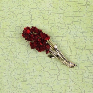 May include: A gold-toned brooch with a cluster of red flower-shaped accents. The brooch is designed to look like a bouquet of flowers.