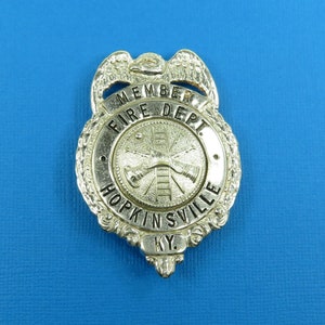 May include: A gold-colored metal badge with the words "MEMBER FIRE DEPT. HOPKINSVILLE KY."  The badge features a stylized image of a firefighter's ladder and hoses.