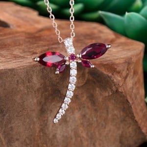 Puede incluir: Un collar con colgante de libélula de oro rosa. La libélula presenta dos gemas ovaladas de color granate para las alas y una fila curva de gemas blancas para el cuerpo. El collar se exhibe sobre una superficie de madera con plantas suculentas verdes en el fondo.