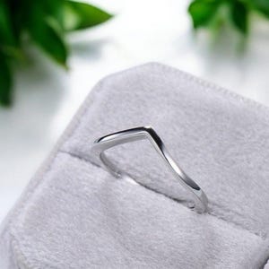 Könnte beinhalten: Ein silberfarbener Ring mit V-förmigem Design, der in einer grauen Samt-Ringbox liegt. Die polierte Oberfläche des Rings reflektiert das Licht, die Box vor einem verschwommenen grünen Hintergrund.