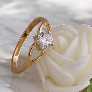 Könnte beinhalten: Ein goldener Ring mit einem großen, runden, klaren Edelstein. Der Ring liegt auf einer weißen Rose mit grünen Blättern. Das Band des Rings hat ein einzigartiges offenes Design.