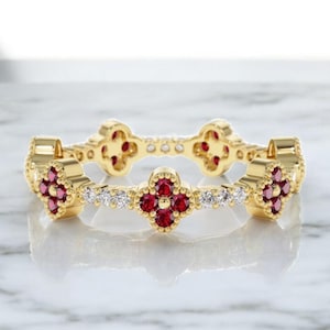 Könnte beinhalten: Ein goldener Ring mit einem sich wiederholenden Kleeblattmuster, besetzt mit roten Edelsteinen und kleinen weißen Diamanten. Die Kleeblattmotive sind gleichmäßig über das Band verteilt. Der Ring liegt auf einer weißen Marmoroberfläche.