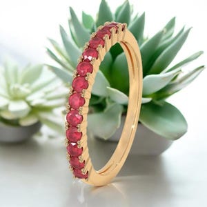 Könnte beinhalten: Ein goldener Bandring mit einer Reihe leuchtend roter Edelsteine. Das Design des Rings zeigt eine durchgehende Reihe gleichmäßig verteilter, rund geschliffener Rubine, die einen funkelnden Effekt erzeugen. Der Ring ist vor einem verschwommenen Hintergrund aus grünen Pflanzen platziert.