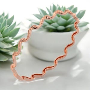Könnte beinhalten: Ein roségoldenes Armband mit einem wellenförmigen, fließenden Design. Die durchgehende Form des Armbands schafft ein einzigartiges Accessoire. Das polierte Metall reflektiert das Licht und verleiht Eleganz. Umfang ca. 18 cm.