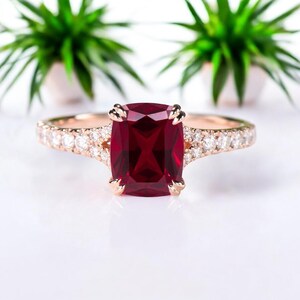 Könnte beinhalten: Ein Ring aus Roségold mit einem großen, tiefroten Edelstein im Kissenschliff in der Mitte, der von vier Krappen gehalten wird. Die Ringschiene ist geteilt und mit einer Reihe kleiner, klarer Steine im Brillantschliff besetzt.