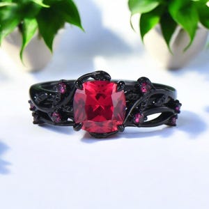Könnte beinhalten: Ein schwarzer Ring mit einem großen, quadratischen, roten Edelstein als Mittelpunkt. Der Ring hat ein florales Design mit kleineren, rosa Edelsteinen. Der Ring ist vor einem weißen Hintergrund mit grünen Pflanzen im Hintergrund platziert.