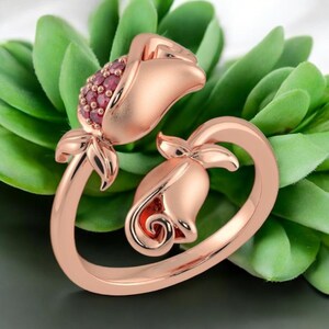 Könnte beinhalten: Roségoldener Ring mit floralem Design, das zwei Rosenblüten zeigt. Eine Blüte ist mit kleinen, roten Edelsteinen verziert. Der Ringbogen ist elegant geschwungen und erzeugt eine romantische Ästhetik. Der Ring ist vor grünem Laub platziert.