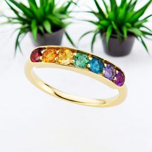 Könnte beinhalten: Ein Goldring mit einer Reihe von sieben rund geschliffenen Edelsteinen in Regenbogenfarben, darunter Rot, Orange, Gelb, Grün, Blau, Indigo und Violett, gefasst in einer polierten Gelbgoldfassung mit einer glatten Oberfläche.