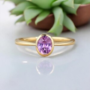 Könnte beinhalten: Ein goldener Ring mit einem hellvioletten, ovalen Edelstein in einer Zargenfassung. Der Ring liegt auf einer weißen Oberfläche, mit einem verschwommenen Hintergrund aus grünen Blättern und einem braunen Topf.