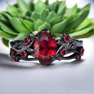 Könnte beinhalten: Schwarzes Ring-Set mit einem großen, oval geschliffenen rubinroten Edelstein und kleineren roten Akzentsteinen. Der Ring hat ein florales Design mit dunklen Metallarbeiten, vor einem verschwommenen grünen Hintergrund.
