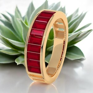 Könnte beinhalten: Ein goldfarbener Ring mit einem Band aus rechteckigen, tiefroten Edelsteinen. Der Ring ist vor einem verschwommenen Hintergrund aus grünen Sukkulentenblättern platziert, was das Design und den Farbkontrast des Schmucks hervorhebt. Das Design des Rings ist modern und elegant.