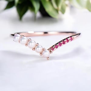 May include: A rose gold ring with a V-shaped design. The ring features a row of white pearls on one side and small red gemstones on the other. The ring is set against a white background.