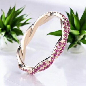 Könnte beinhalten: Ein goldfarbener Ring mit einem gedrehten Design, besetzt mit kleinen, leuchtend rosa Edelsteinen. Der Ring ist vor einem weichen, weißen Hintergrund platziert, mit grünen Pflanzen im Hintergrund. Das Design des Rings ist elegant und auffällig.