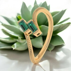 Könnte beinhalten: Ein goldfarbener Ring mit zwei rechteckigen, smaragdgrünen Edelsteinen. Der Ring hat ein einzigartiges, geschwungenes Design mit einem strukturierten Band und einer glatten, polierten Oberfläche. Die Edelsteine sind in einer goldenen Lünette gefasst.
