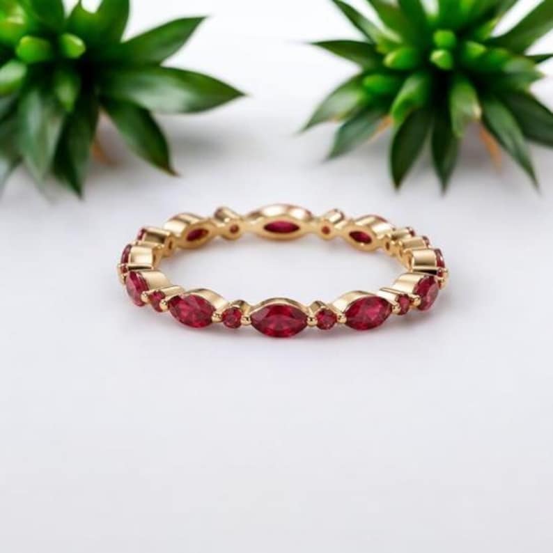 Puede incluir: Un anillo de banda dorada con un patr&oacute;n alterno de piedras preciosas rojas de corte marquesa y corte redondo. El anillo est&aacute; construido con un acabado de metal pulido, mostrando un dise&ntilde;o continuo de piedras facetadas engastadas en el marco de oro.