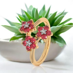 Könnte beinhalten: Ein goldener Ring mit drei Blumendesigns. Jede Blume besteht aus roten Edelsteinen und einem zentralen Cluster aus kleinen weißen Edelsteinen. Der Ring ist vor einem verschwommenen Hintergrund aus grünem Laub angeordnet.