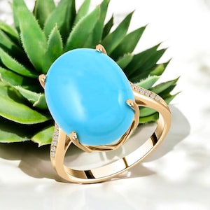 May include: A gold ring featuring a large, oval, light blue gemstone. The ring's band is gold, with small accent stones set along the sides of the gemstone. The ring is set against a background of green foliage.