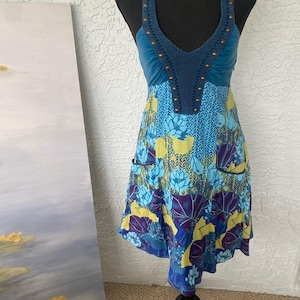 May include: A blue and yellow floral print dress with a halter top. The top is a dark blue crochet with bronze-colored studs. The dress has a V-neck and two small pockets. The dress is displayed on a black mannequin.